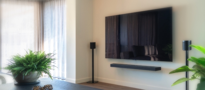 Stue med hvide gardiner til venstre og et fjernsyn med soundbar og to separate højttalere på højttalerstandere til højre på væggen